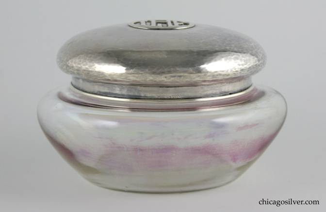 Kalo jar for powder, two-piece, with glass bottom and separate sterling top.  Jar is hand-blown rainbow glass with flat bottom, flaring sides, cylindrical neck.  Tight-fitting silver lid is mushroom shaped, with some top centering an applied LHF mono inside a circle.  Bottom of the lid has applied wire around the rim.  Fairly heavy, and nice hammering.  Top:  4-3/8" W and 1-1/4" H.  Glass bottom:  5-1/4" W and 2-1/4" H.  Together:  5-1/4" W and 3-1/4" H.  Marked:  STERLING KALO
