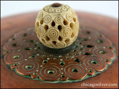 Detail from Rebecca Cauman bowl, round, copper, with removable lid, white enameled interior and delicately carved and pierced spherical ivory finial on lid atop centered decoration of applied chased concentric circles with pierced small circles and teardrop shapes.  