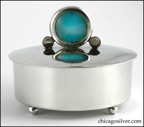 Rebecca Cauman box, round, pewter, straight-sided on flat bottom with four small ball feet, and removable tightly fitted slightly domed lid.  Lid has large upright round translucent blue stone finial in round see-through frame with a small solid round disk on either side, and eight chased lines radiating outward from the center.  