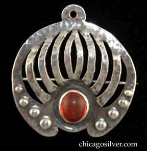 Forest Craft Guild brooch, round, German silver, with small tab at top with hole for chain, plus pin with safety clasp at back, nine grill-like curving cutouts below the tab, several graduated repousse domes on bottom edges, and large oval bezel-set red cabochon stone at bottom center.  