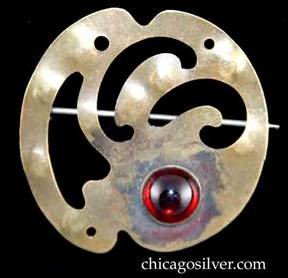 Forest Craft Guild brooch, round, brass, with notches top and bottom, three curving cutouts at center, three pierced holes around edge, and round red bezel-set cabochon stone at bottom. 