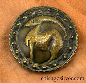 Forest Craft Guild brooch with original tan ooze leather backing.  Brass, round, with dark patina, wide circular decoration on edge of two concentric rings with 18 repouss dimples between them in mushroom-shaped forms, centering a lifelike rendering of a full-body ibex, with long notched horns and arched back.  Brooch is pinned to original brown ooze leather presentation material.  Hammered edges.
