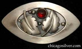Foss brooch, oval, with pointed ends, wide raised edge, two applied serrated leaves with curving central vein, silver beads, oxidized background, and round red bezel-set garnet stone.  