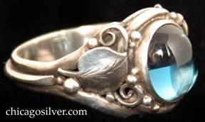 Laurence Foss ring, silver, delicate, with oval bezel-set cabochon light blue topaz stone set at a slight angle in oxidized frame surrounded by a smooth applied leaf with chased details and beads and wirework decoration on each side.  A wire runs around the back of the ring connecting the bottoms of each leaf stem.  