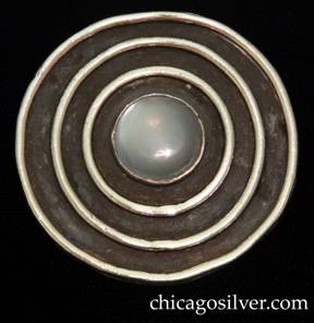 Mary Gage brooch / pin, circular frame with open back and three concentric thick rings on front centering a bezel-set cabochon moonstone.  Outermost circle is somewhat irregular.  Heavy.  