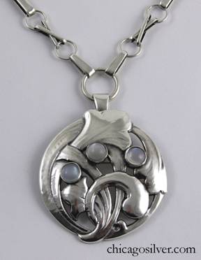 Kalo necklace / brooch, very large round pendant with pierced frame containing stylized chased floral elements that curve around and through each other and the frame, with a series of three round bezel-set cabochon moonstones.  Back is flat.  Folding bale and pin on back for use as brooch.  Unusually heavy chain (the kind seen on lockets) with spring ring at bottom to remove it from pendant bale, and large wide oval loops alternating with pairs of thin oval loops in a x-shaped arrangement, connected to one another with wide circular loops.  