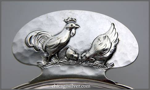 Kalo porringer, round, flat bottom form with rounded tab handle and applied wire to outside of rim.  Handle has chased and repoussé decoration of rooster standing on grass beside hen and chick pecking at ground -- detail shown here