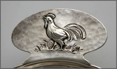 Kalo porringer, round, flat bottom form with tab handle and applied wire rim.  Handle has chased and repoussé decoration of rooster standing on grass.  Detail shown.