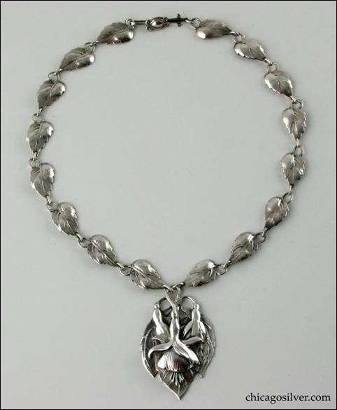 Necklace, silver, composed of 16 leaf-form links each with a stem that curves around and forms a loop, centering a large pointed oval plaque with chased fuchsia blossoms and leaves.  Plaque is heavy and dimensional.  Long thin interesting clasp with applied curving decoration.